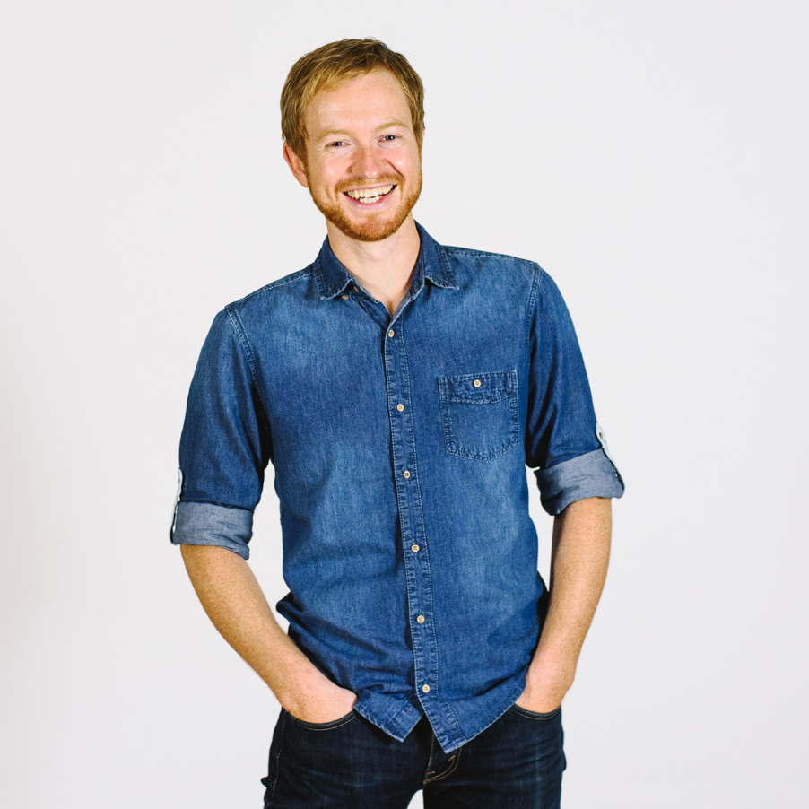 Ben White smiling at the camera, wearing a blue denim shirt, standing against a white background